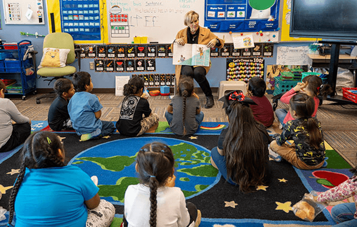 Teacher reading a book to a classroom of students sitting on the floor. 