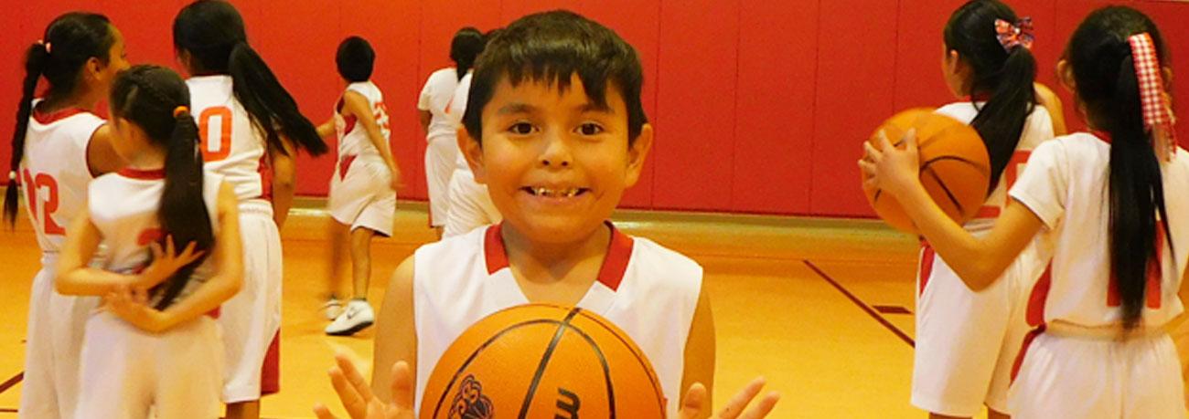 A smiling young basketball player holds a basketball in the foreground as children in white and red uniforms practice on an indoor basketball court.