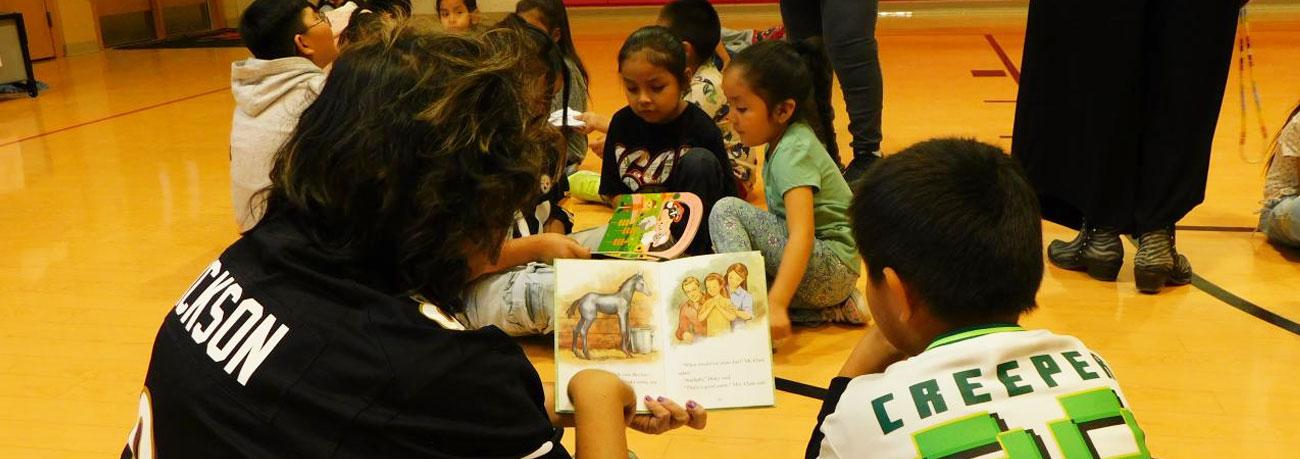  Children sitting on an indoor basketball court, looking at books and reading together while adults supervise nearby.